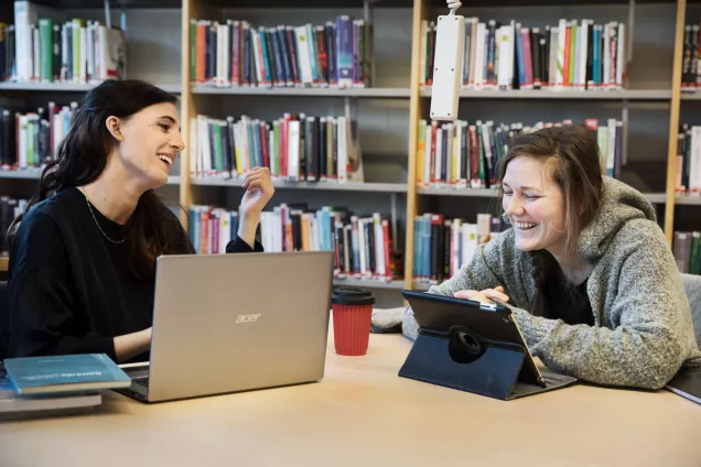 Two students laughing in a library with a laptop and a tablet